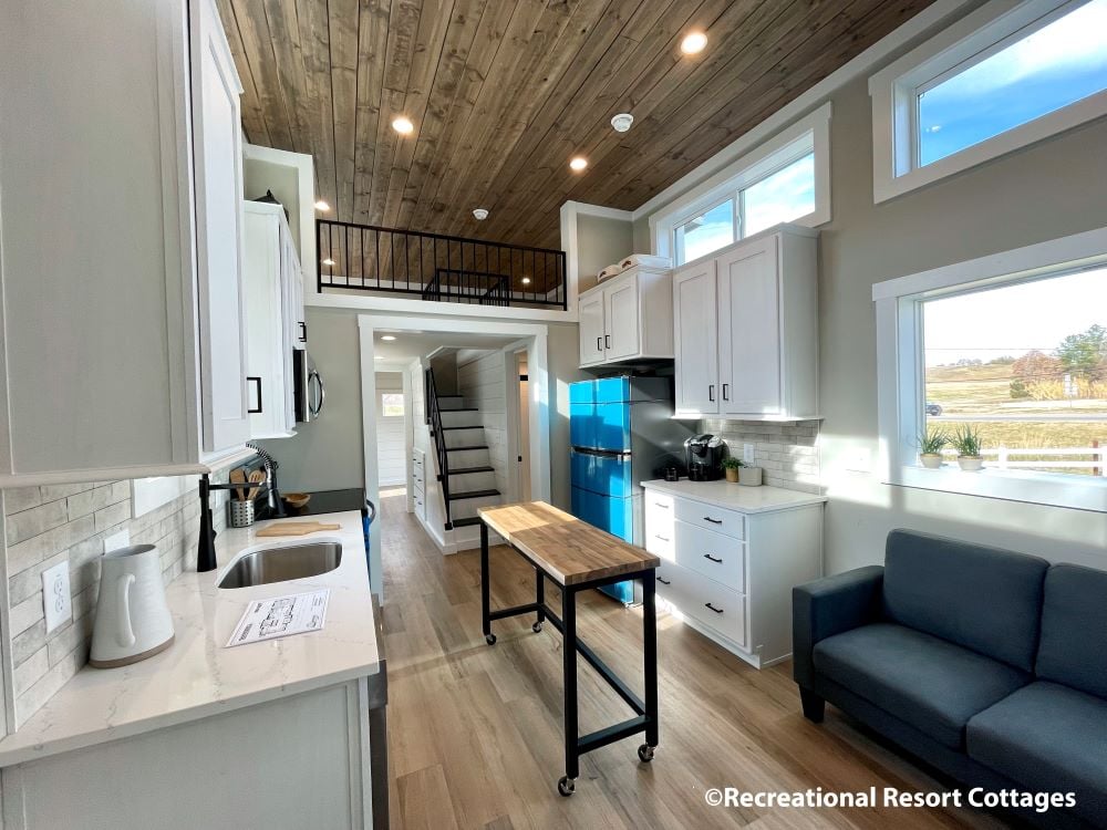 Modern tiny house interior with a wood ceiling and floor. Features include white cabinets, a central island, blue fridge, cozy sofa, and a loft with a metal railing. Bright windows provide natural light, creating a warm atmosphere.