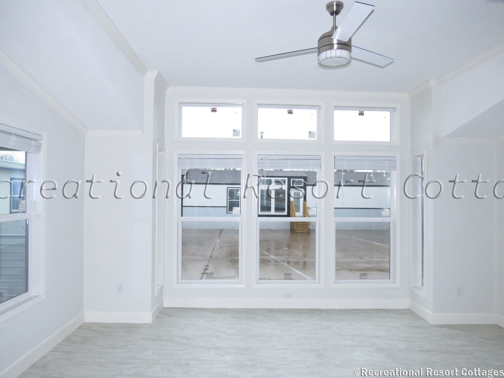 Bright, minimalist white room with large windows, tiled floor, and modern ceiling fan. The space feels open and airy, looking out onto a patio.
