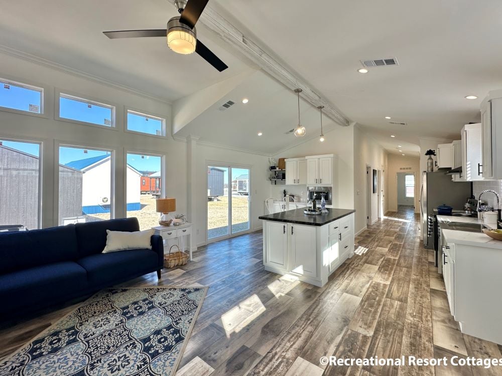 Spacious, sunlit living area with high ceiling, white cabinets, and hardwood floors. Blue sofa and patterned rug add cozy touches. Modern and inviting.