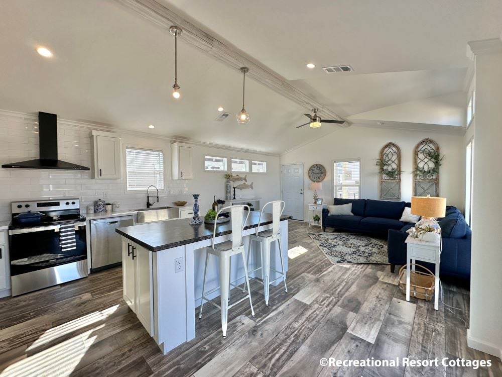 Spacious open-concept living room and kitchen with vaulted ceilings. Features white cabinetry, wood floors, a navy sofa, and modern decor. Bright and inviting.