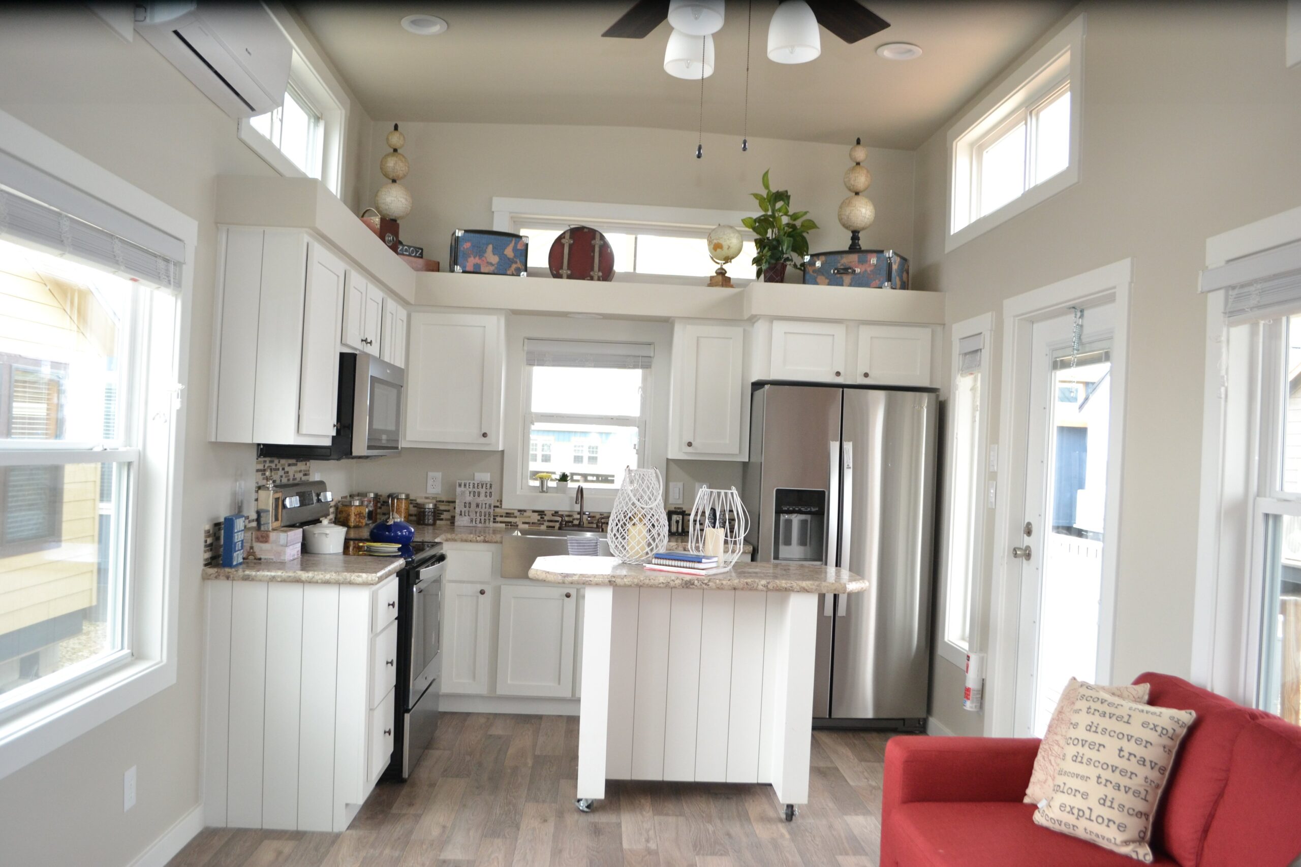 A cozy kitchen with white cabinets, stainless steel appliances, and a central island. Decor includes plants and ornaments, creating a welcoming and airy atmosphere. A red couch with a decorative pillow adds a pop of color.