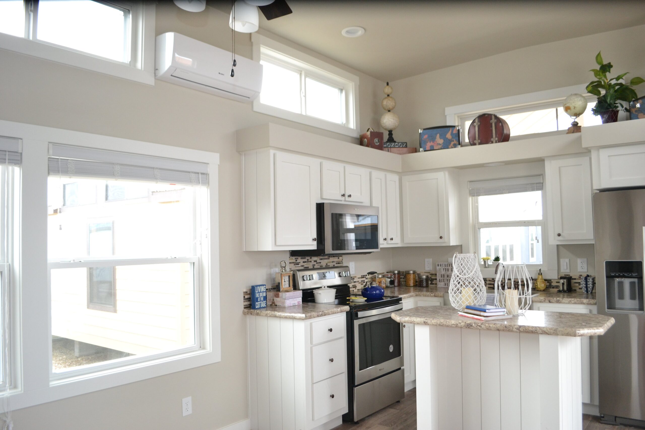 Bright kitchen with white cabinets, stainless steel appliances, and a small island. Sunlight streams through large windows, creating a cozy atmosphere.