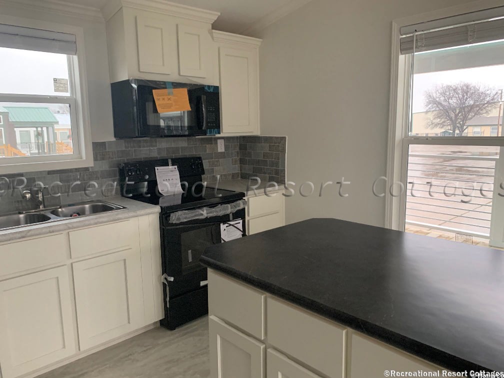 A compact kitchen features white cabinets, a black countertop, and appliances. Large windows reveal an overcast day, creating a cozy ambiance.