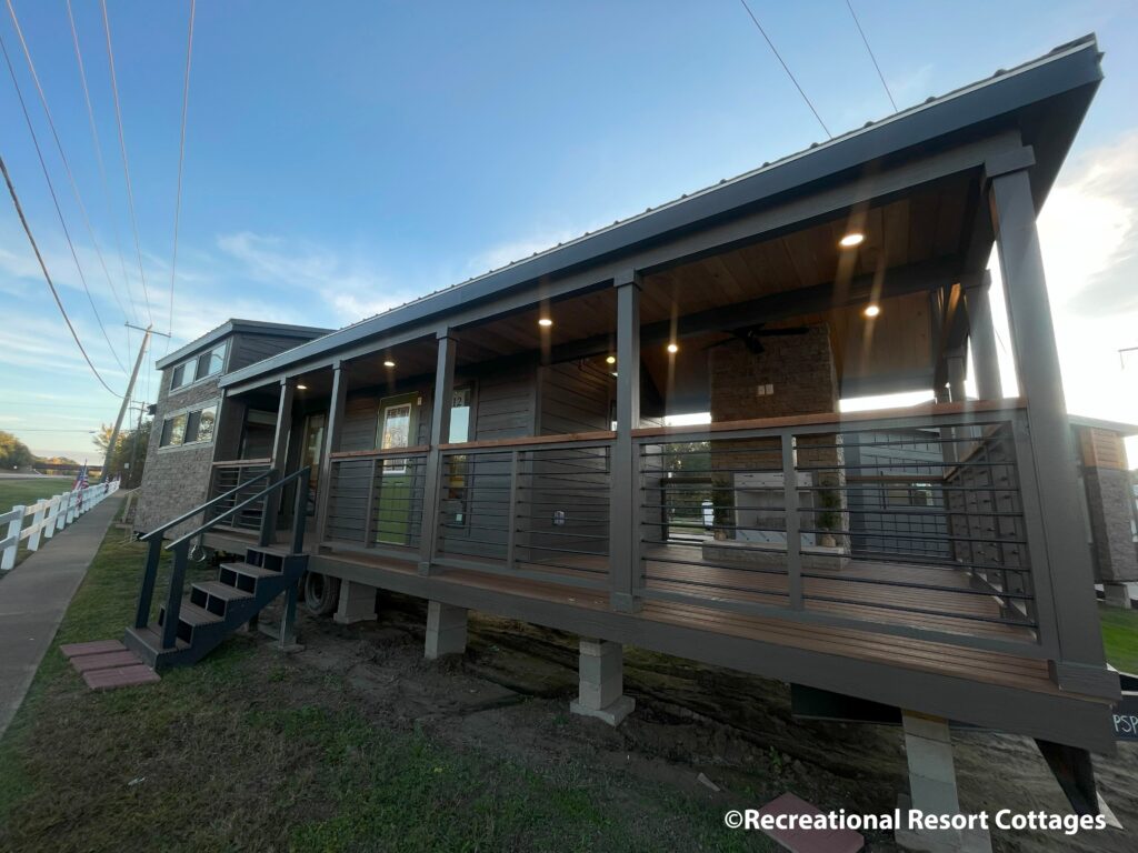 Modern tiny house with a covered porch and steps, set at dusk. Warm lights illuminate the wooden facade, creating a cozy atmosphere. Grass and sidewalk nearby.