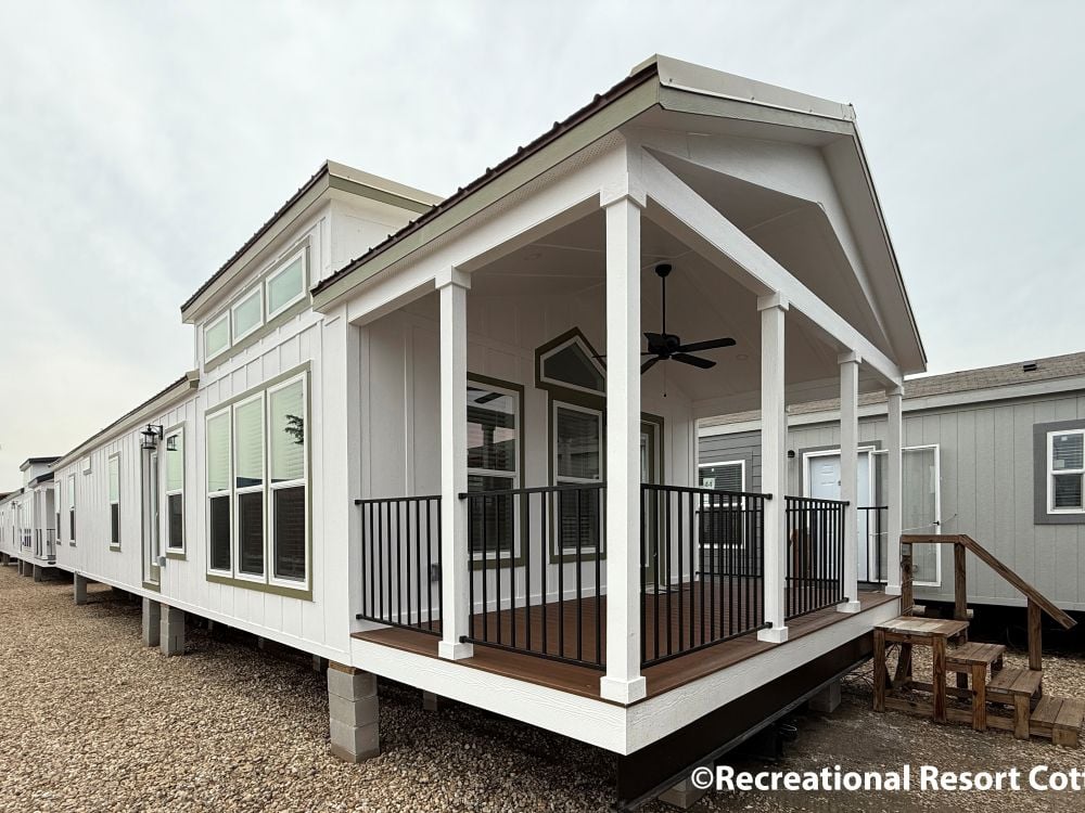 Modern white manufactured home with a spacious covered porch featuring railing and ceiling fan. Large windows enhance its contemporary design.