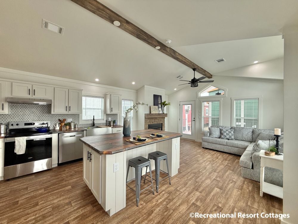 Spacious open-plan kitchen and living area with modern decor. Features wood floors, a large island with barstools, stainless steel appliances, and a cozy gray sofa.