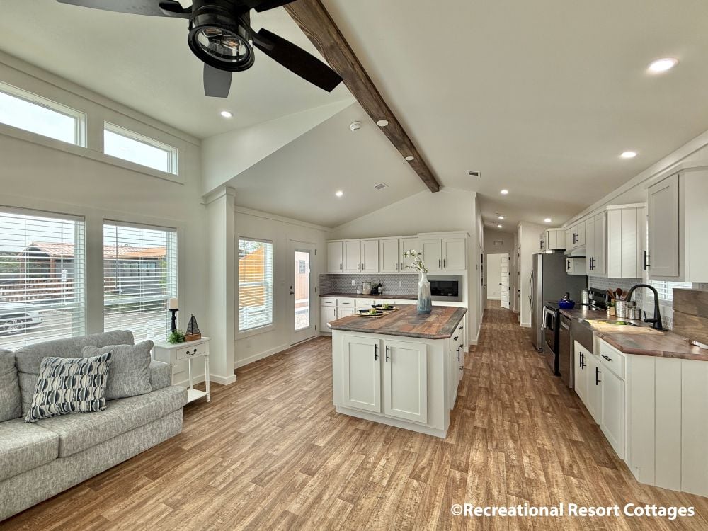 Spacious modern kitchen and living area with wood floors, white cabinets, and a central island. Large windows and a cozy gray couch enhance the airy feel.