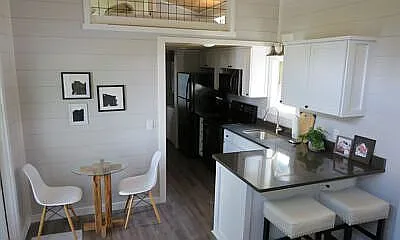 Compact white kitchen with modern black appliances, a small dining area with two chairs and a round table, and a cozy breakfast bar with stools. Minimalist decor adds charm.