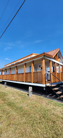 A quaint white house with a red-roof, wooden porch, and railings stands under a clear blue sky. It exudes a warm, welcoming atmosphere. Grass surrounds the elevated structure.