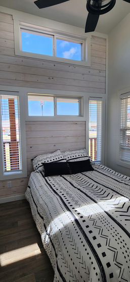 Cozy bedroom with a bohemian-patterned bedspread, wooden accent wall, and large windows letting in natural light, creating a bright, airy atmosphere.