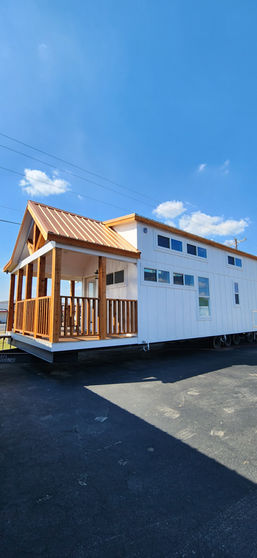 A small white house with a wooden front porch and a copper-colored roof stands on a paved area. The sky is clear with a few clouds, conveying a serene atmosphere.