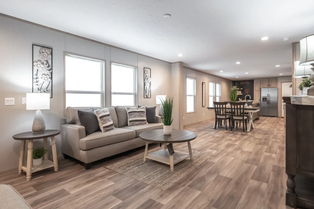 Modern open-concept living room and kitchen. Features a gray sofa with cushions, round wooden table, dining set, and sleek kitchen, all in neutral tones.