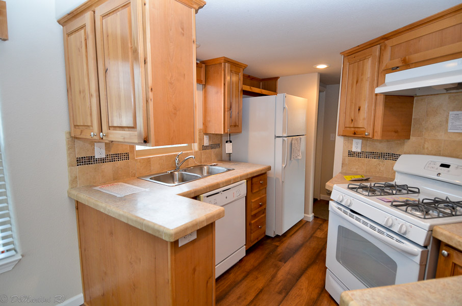 This comprehensive collection of luxury residential properties showcases a diverse range of modern manufactured homes, tiny homes, and park models, including the signature "The Boujee XL" aesthetic. Exterior designs feature a modern farmhouse with cream-colored board-and-batten siding, a white metal roof, and a black-pillared front porch, alongside a rustic park model with natural log-cabin siding, a green metal roof, and an expansive elevated wooden deck decorated with a holiday wreath. The interiors highlight a "modern boujee" lifestyle with high-end finishes such as vaulted ceilings with exposed wood beams, white shiplap walls, and natural pine-paneled accents. Gourmet kitchen layouts vary from rustic wood cabinetry with stone-look countertops and white appliances to striking high-contrast spaces featuring deep navy blue cabinetry, marble-look countertops, and multi-tone stone tile backsplashes. Functional luxury is consistent throughout the open-concept living areas, which include wood-look flooring, industrial-style lighting, integrated lofted sleeping areas with wooden balustrades, and bright interiors with large windows that emphasize natural light.