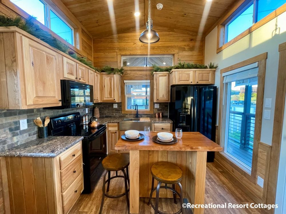 Cozy wooden kitchen with vaulted ceiling, illuminated by natural light. There’s a central island with two stools, set with plates and glasses, conveying warmth.