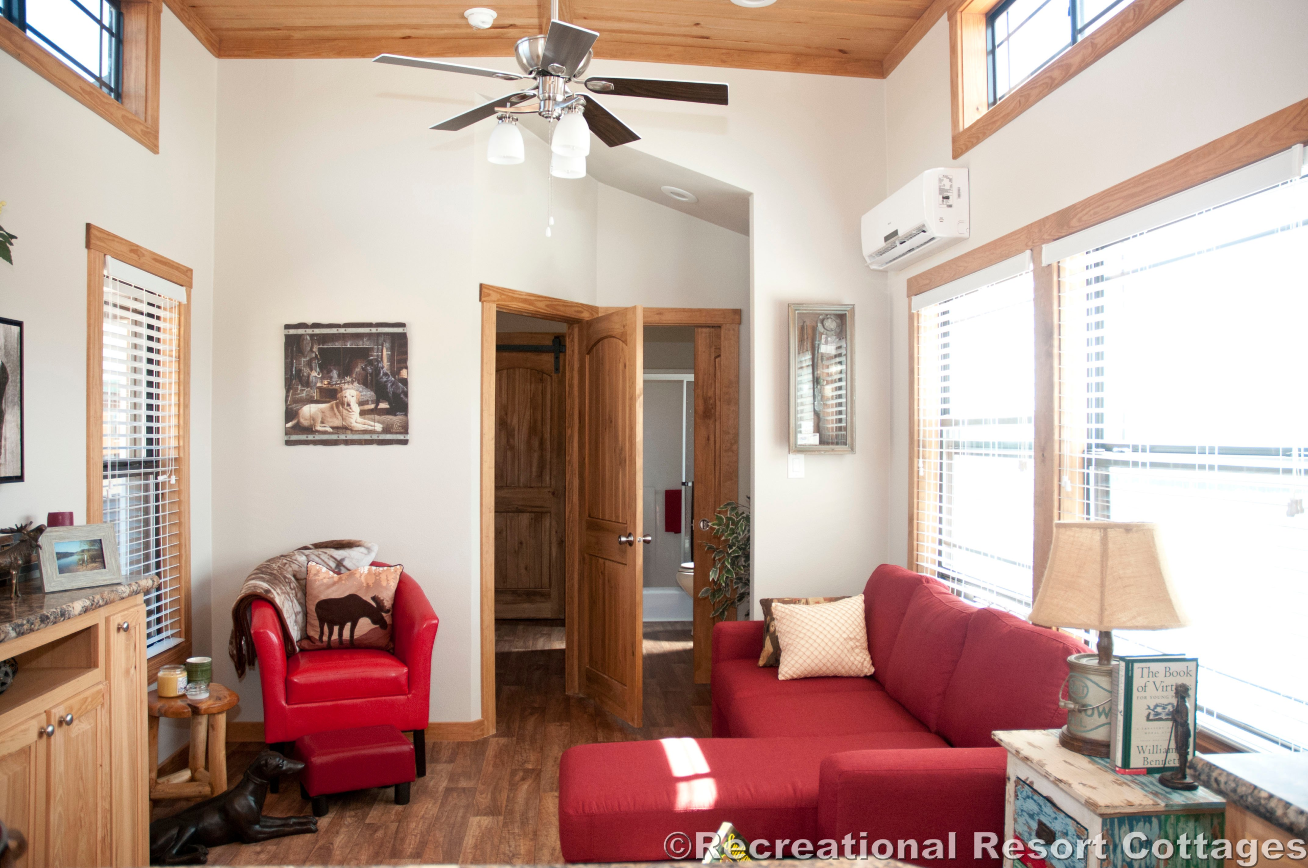 Cozy living room with a red sofa, armchair, wooden floors, and ceiling fan. Sunlight streams through large windows, creating a warm, inviting atmosphere.