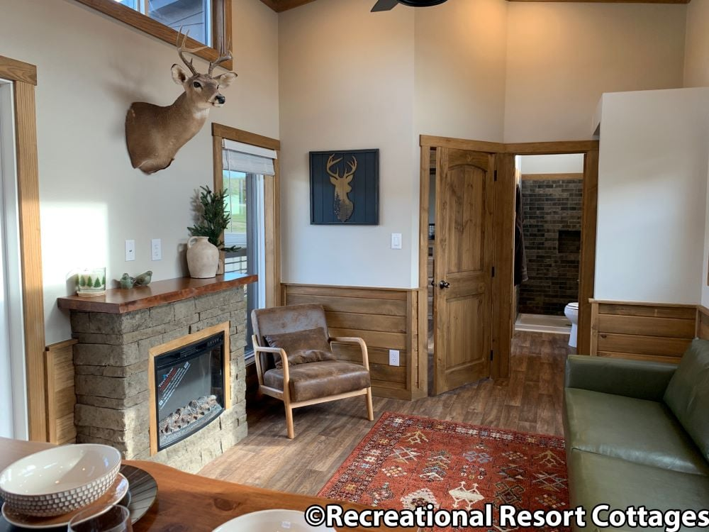 Cozy cottage living room with wood accents. Features a mounted deer head, stone fireplace, green sofa, leather chair, and a rustic red rug. Warm ambiance.