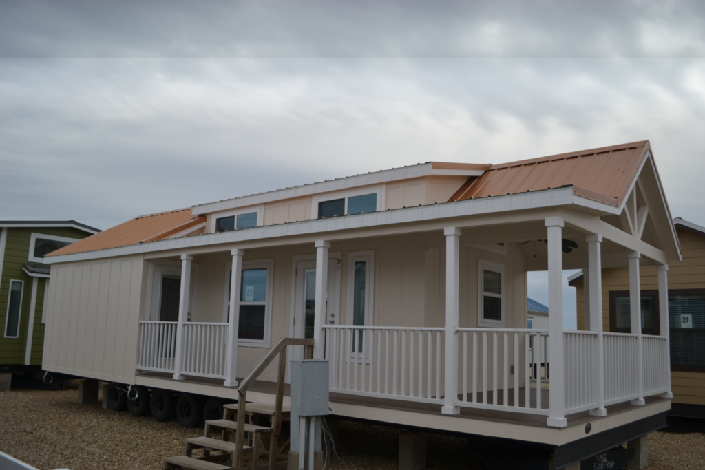 A beige modular home with a white railing porch and orange metal roof sits on a gravel lot under a cloudy sky, conveying a sense of simplicity and modernity.