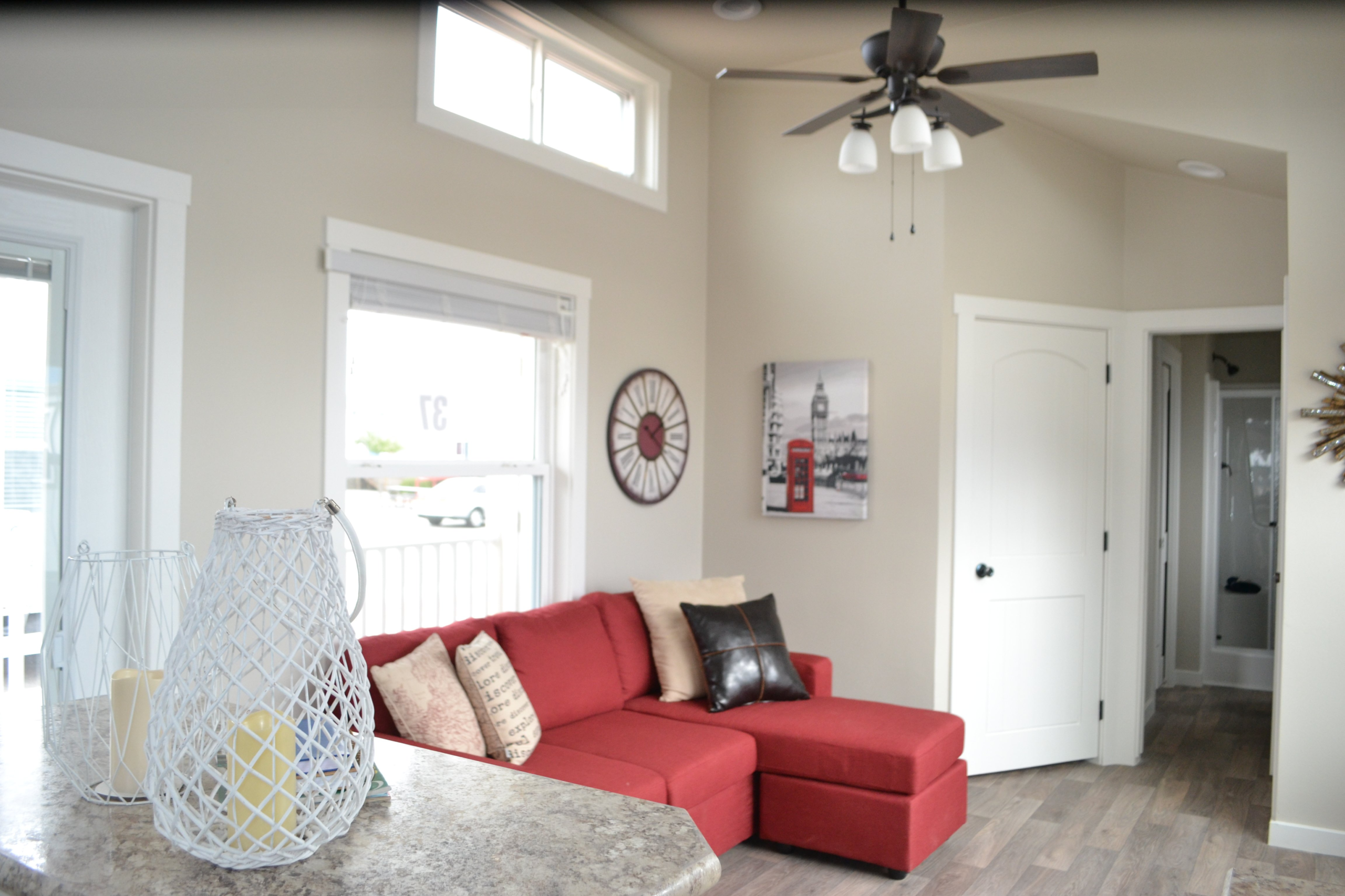 A cozy living room with a red sectional sofa and accent pillows against beige walls. A wall clock and London-themed artwork add charm. A lighted ceiling fan and tall windows create an airy feel. A marble countertop holds decorative white candleholders.