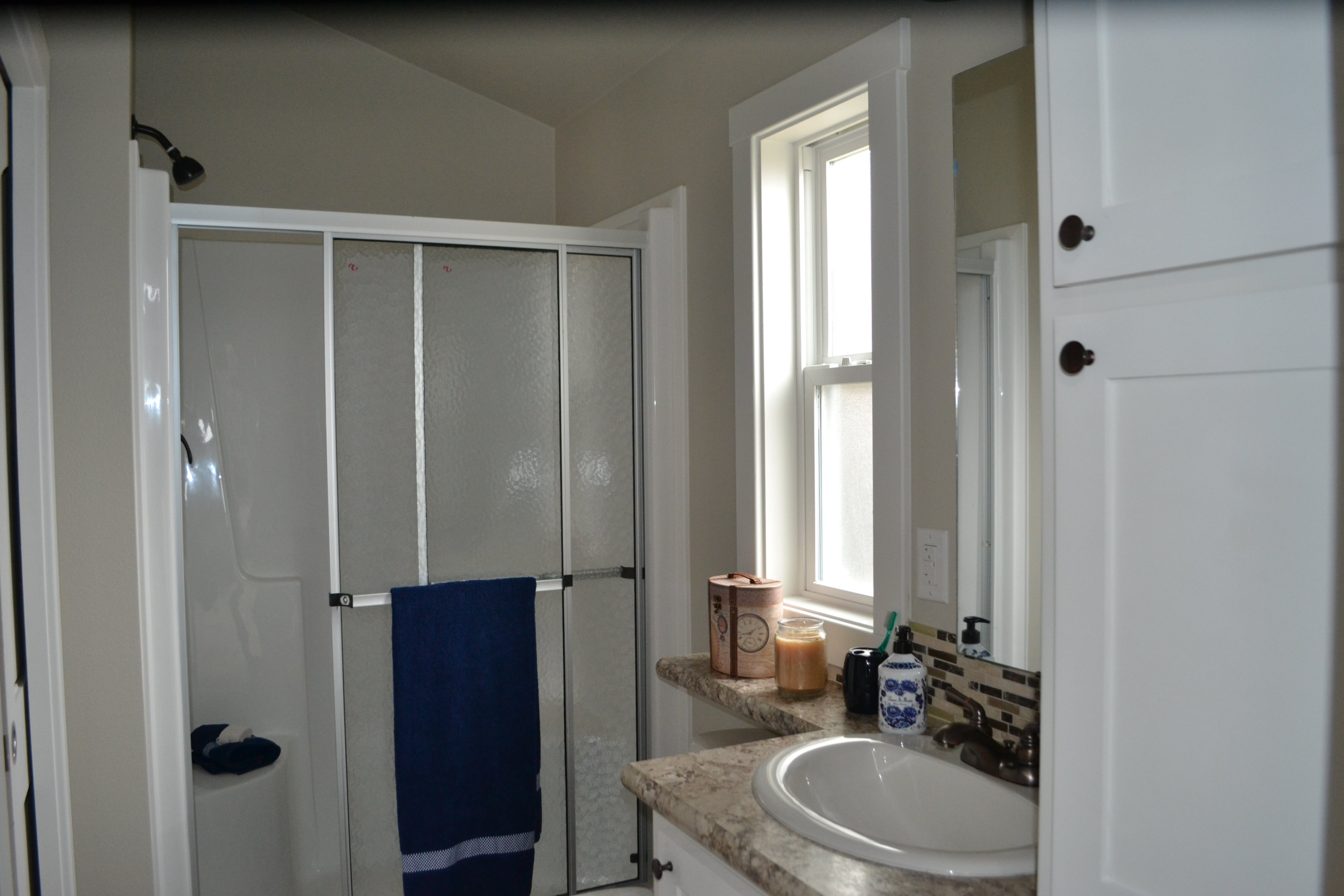 A clean bathroom with a glass shower door, blue towel, and beige walls. A sink with toiletries, a window, and cabinetry add to the bright, tidy space.
