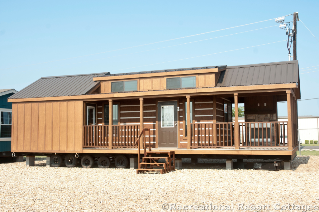 A rustic, brown tiny house with a metal roof and a porch sits on gravel. Wooden steps lead to the entrance, conveying a cozy and inviting atmosphere.