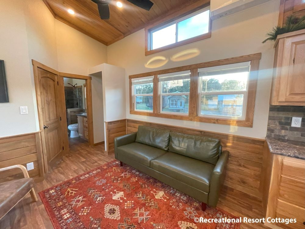 Cozy room with wood paneling and vaulted ceiling. Green leather sofa faces large windows, reflecting soft daylight. Red patterned rug adds warmth.