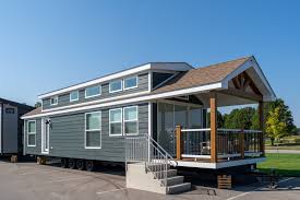 A modern tiny house with a gray exterior and white trim, featuring large windows, a gabled roof, and a small porch. It is set in a sunny outdoor scene.
