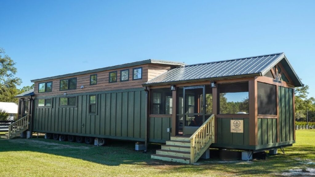 A modern tiny house on wheels, featuring green siding, a metal roof, and a raised porch with stairs. Set against a clear blue sky, it conveys a cozy, inviting atmosphere.