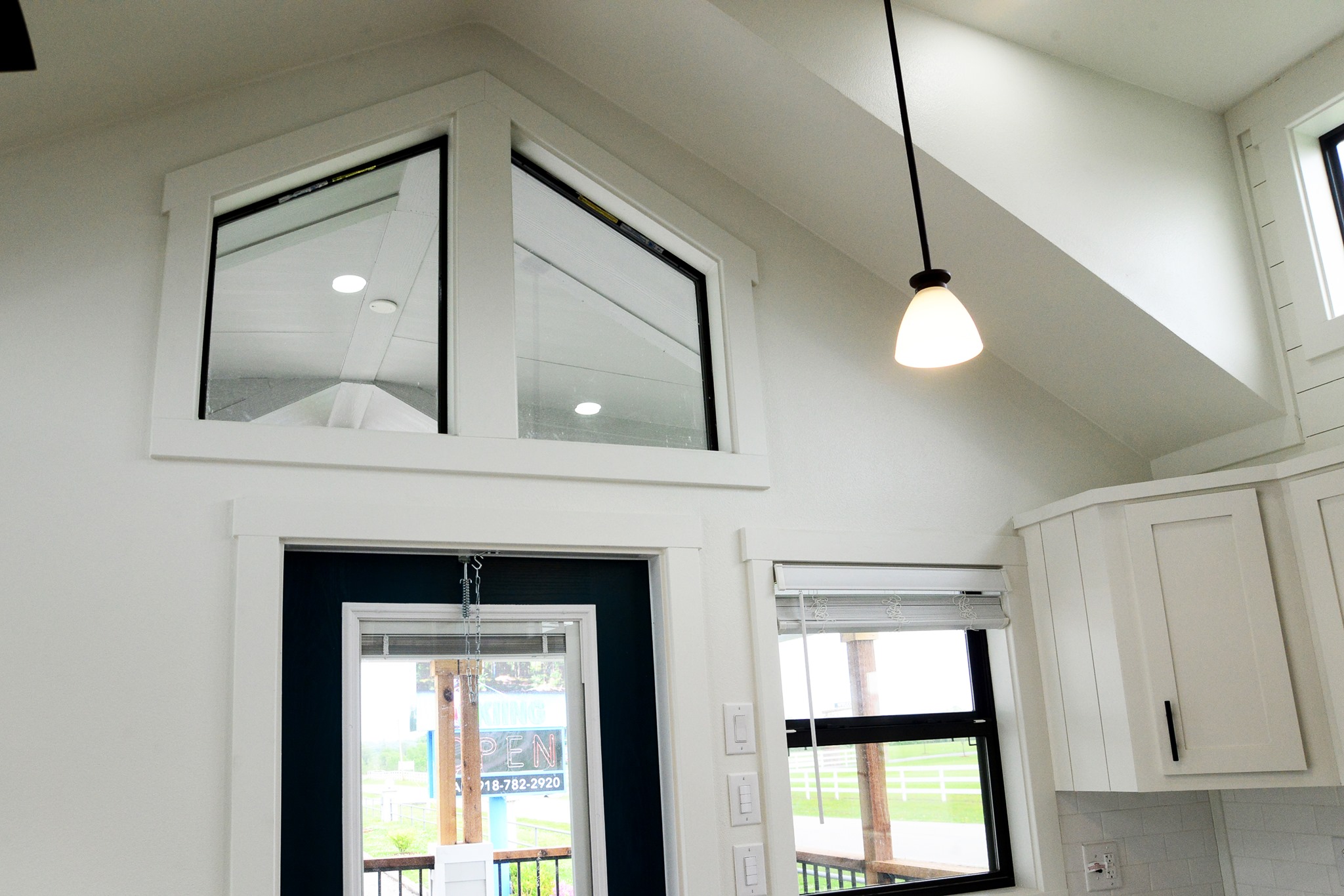 Interior of a modern tiny house featuring a vaulted white ceiling with a pendant light, a dark framed triangular window above a dark blue door with a glass pane showing an "OPEN" sign, and a white kitchen cabinet with a black handle next to a window with white blinds, all under bright natural light.