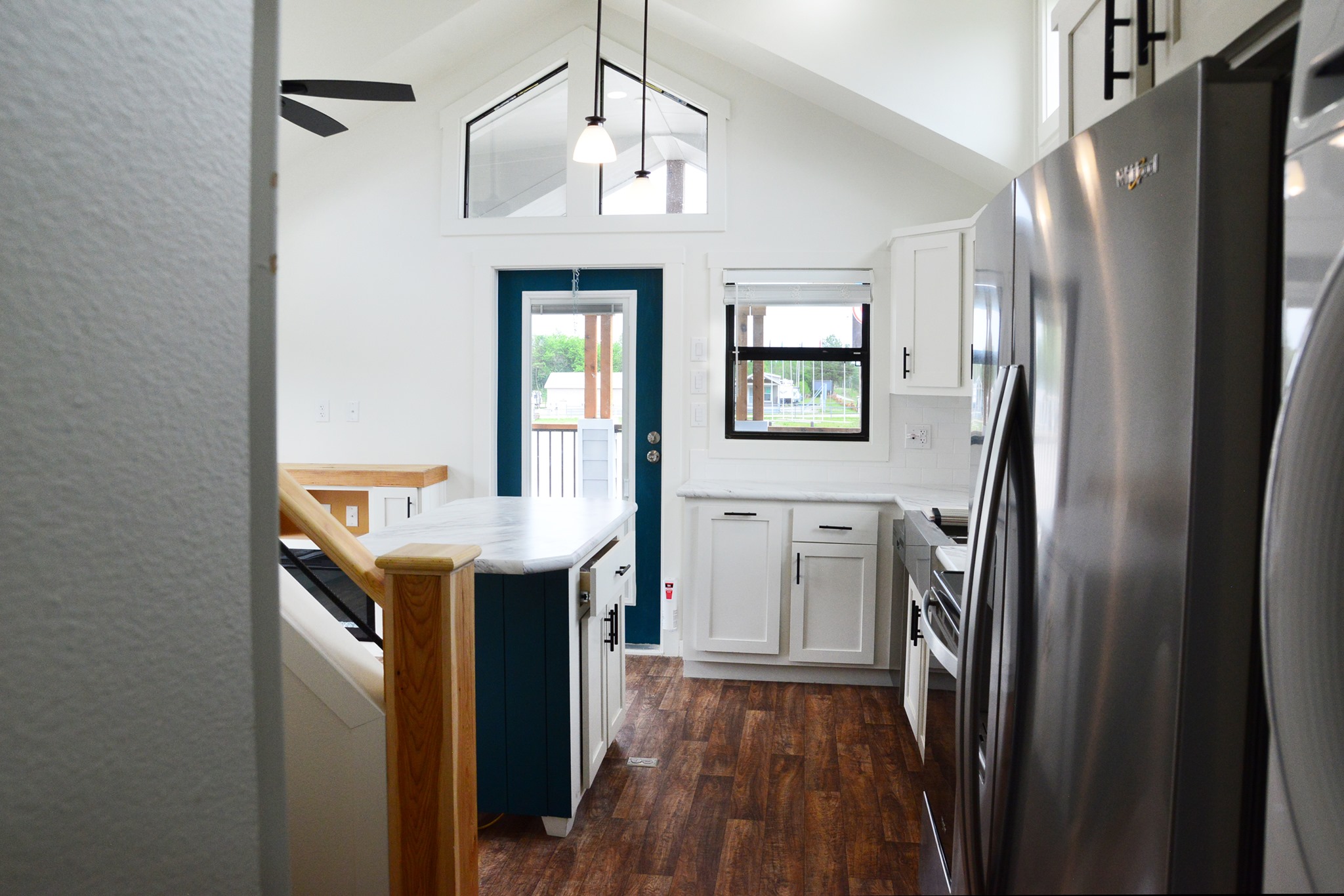 Interior view of a modern tiny house kitchen with white shaker cabinets, marble countertops, a dark teal island, stainless steel appliances including a refrigerator and stovetop, a blue front door with a transom window, and wood-look flooring under a vaulted ceiling with a ceiling fan and pendant lights, offering a glimpse of a bright and functional living space.