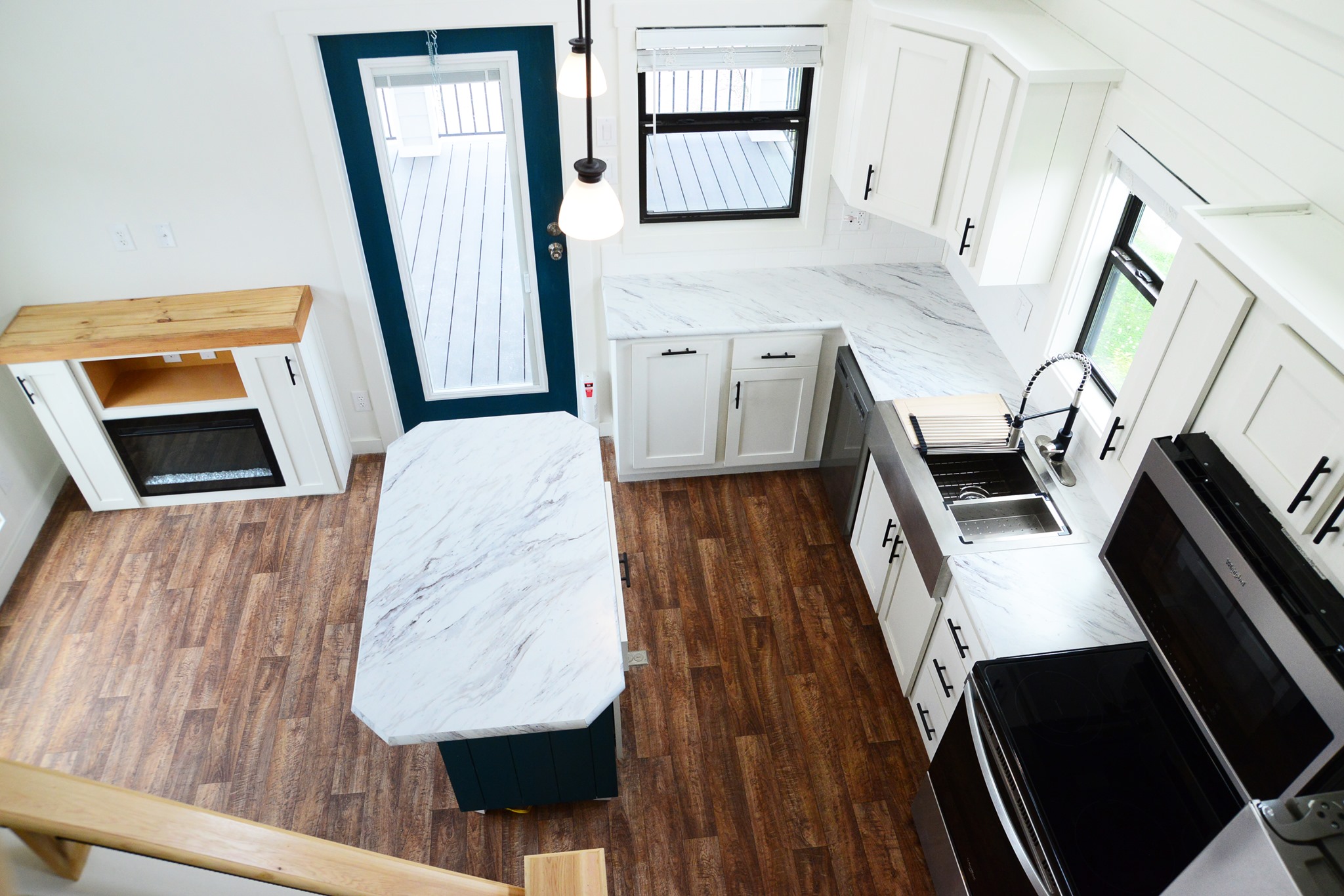 Interior view of a modern tiny house showcasing a bright white kitchen with marble-patterned countertops, white shaker cabinets with black hardware, a stainless steel farmhouse sink with a pull-down faucet, a black electric stovetop, and a stainless steel microwave, complemented by a dark teal kitchen island with a matching countertop and seating, a cozy electric fireplace with a wooden mantel, and wood-look laminate flooring, all viewed from an elevated angle looking down into the compact and stylish living space.
