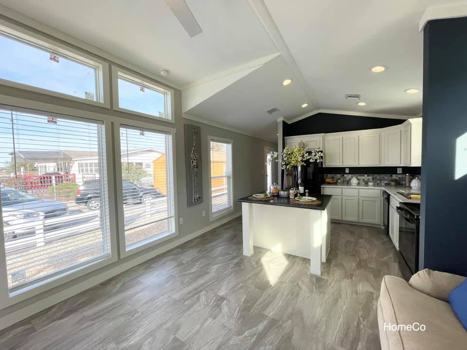 Modern kitchen with large windows, white cabinets, and a black countertop. Sunlight floods the space, highlighting a vase with flowers. Cozy and inviting.