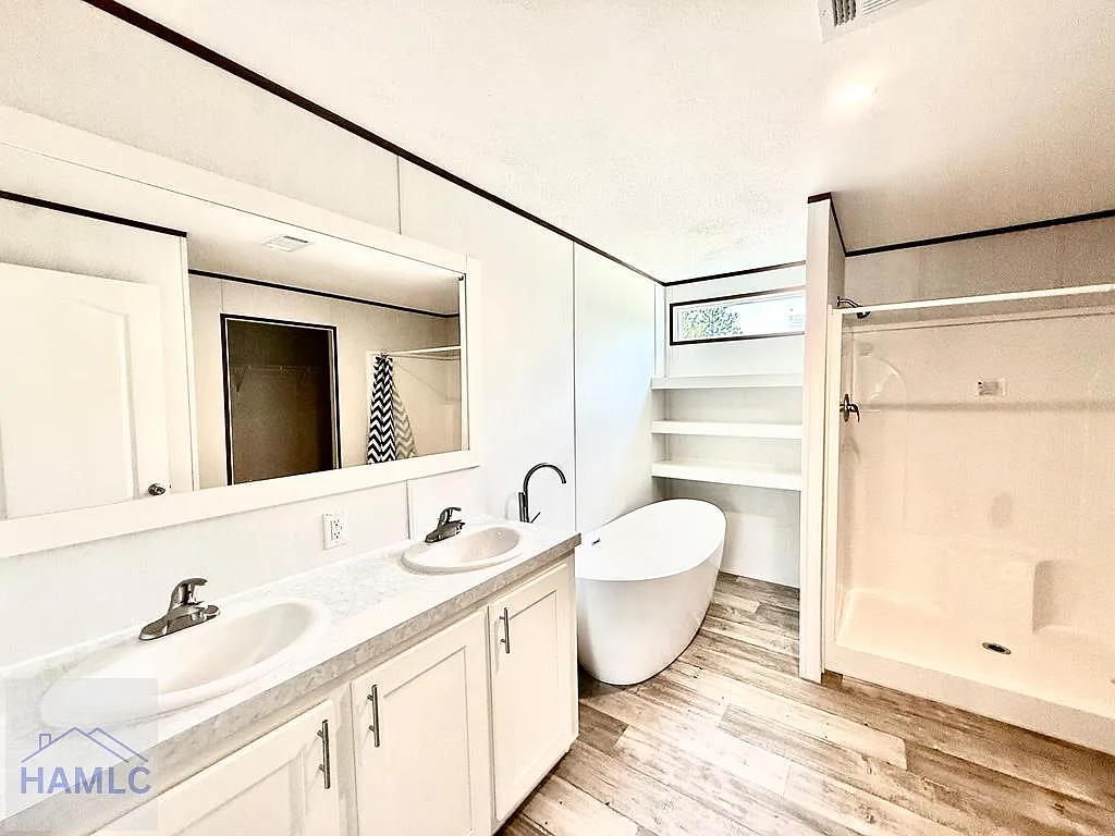 Luxurious modern primary bathroom in premium manufactured home featuring white shaker vanity cabinets with brushed nickel hardware dual undermount sinks matte black faucets large framed mirror with integrated lighting white freestanding oval soaking tub spacious walk-in shower with seamless glass enclosure built-in shelving niche gray wood-look luxury vinyl plank flooring light gray walls with dark trim accent tray ceiling recessed lighting small window for natural light bright spa-like ensuite master bath interior design 2025 double-wide modular home by HAMLC