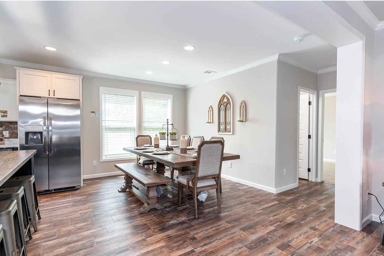 Stunning farmhouse dining room featuring a rustic reclaimed wood dining table with a matching bench and upholstered chairs, set against a backdrop of large windows with white blinds, a stainless steel refrigerator and white cabinetry in the adjacent kitchen area, and decorative arched wall decor, all unified by distressed wood-look laminate flooring and a neutral color palette.