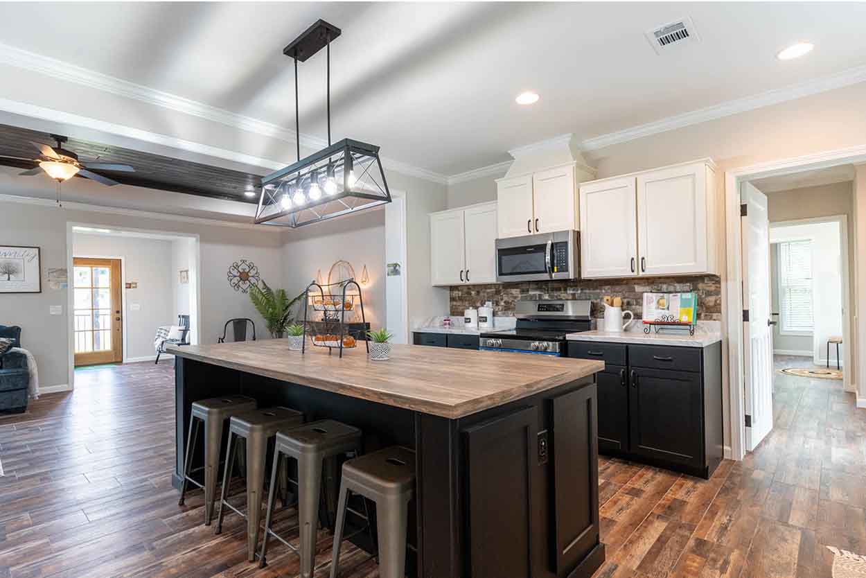 Spacious open-plan kitchen and living area in a manufactured home, featuring a large dark-paneled island with a light wood countertop and four metallic stools, a modern black and white kitchen with stainless steel appliances, white upper cabinets, dark lower cabinets, and a brick-patterned backsplash, all illuminated by a stylish geometric pendant light and recessed lighting, with distressed wood-look flooring unifying the space and a glimpse of a living room with a navy sofa and a wooden door in the background.