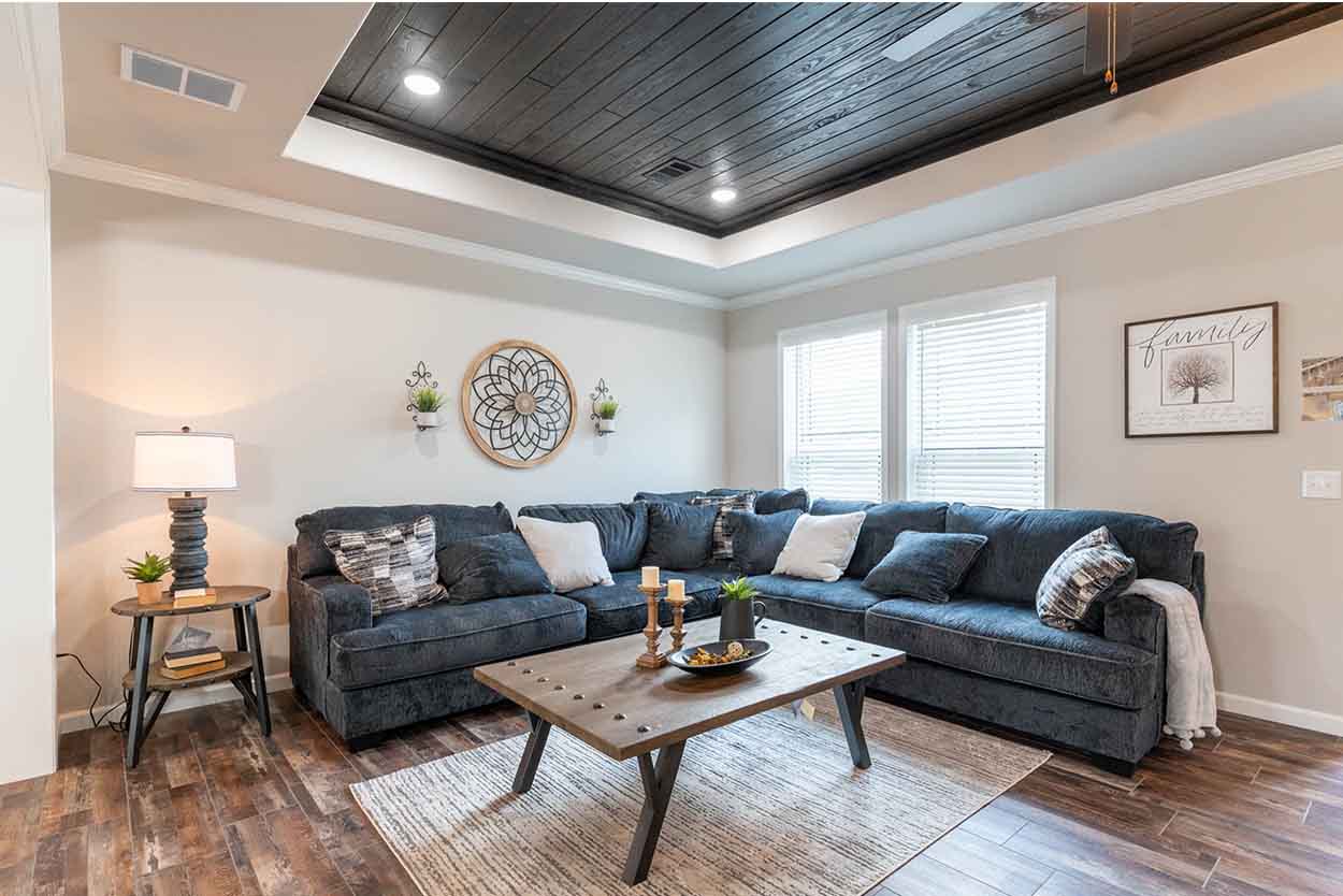 Spacious living room in a manufactured home featuring a large, comfortable navy sectional sofa adorned with decorative pillows, a rustic wooden coffee table with metal accents, and a textured area rug, all under a dramatic dark wood-paneled tray ceiling with recessed lighting and a stylish ceiling fan, complemented by light-colored walls with a large circular wall decor and framed art, and wide windows with blinds providing natural light, all set on a distressed wood-look laminate floor.