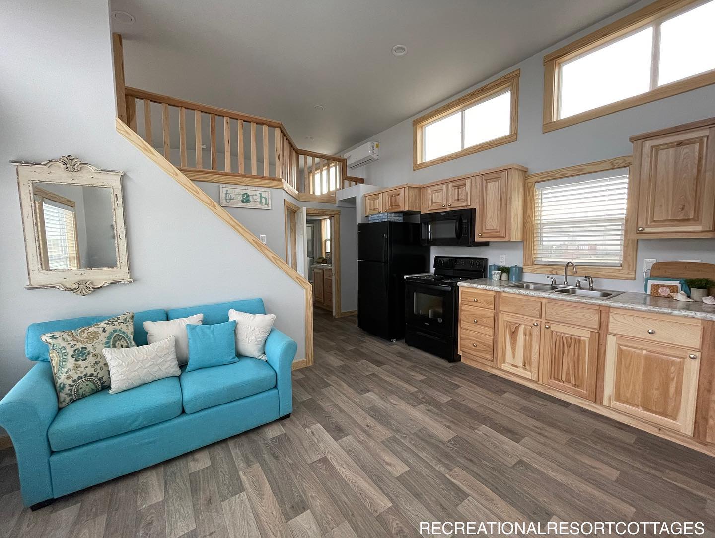 Modern luxury tiny home interior featuring a vibrant blue sofa with decorative pillows, a rustic galley kitchen with natural wood cabinetry and black appliances, and a staircase with a wooden railing leading to a cozy loft area, all set on premium grey wood-look flooring with large clerestory windows for a bright and sophisticated move-in ready turn-key residential design.