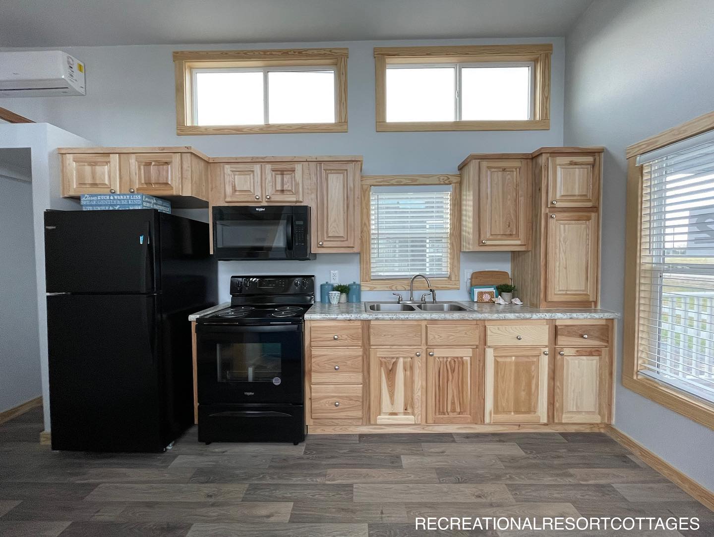 Modern luxury tiny home interior featuring a vibrant turquoise sofa with decorative pillows, a rustic galley kitchen with natural wood cabinetry and black appliances, and a staircase with a wooden railing leading to a cozy loft area, all set on premium grey wood-look flooring with large clerestory windows and a vintage-style ornate mirror for a bright and sophisticated move-in ready turn-key residential design.