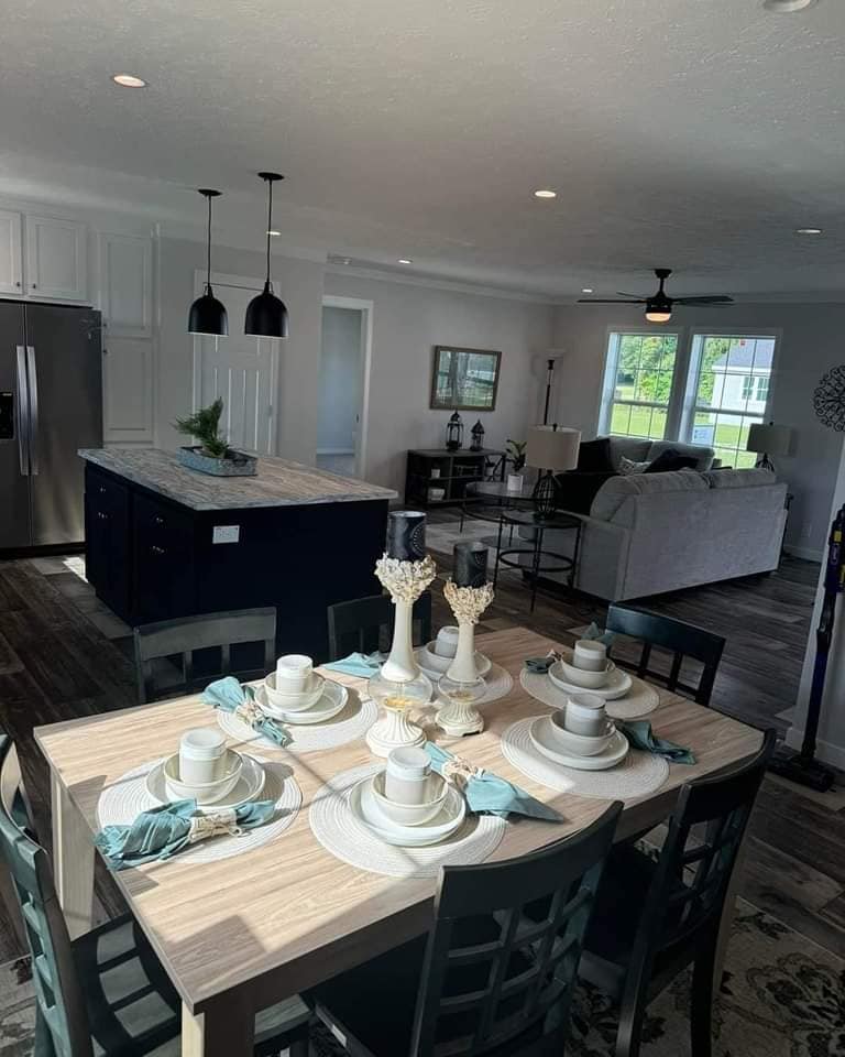 Dining and living area with a wooden table set for six, featuring white tableware and turquoise napkins. Behind is a modern kitchen and cozy living room.