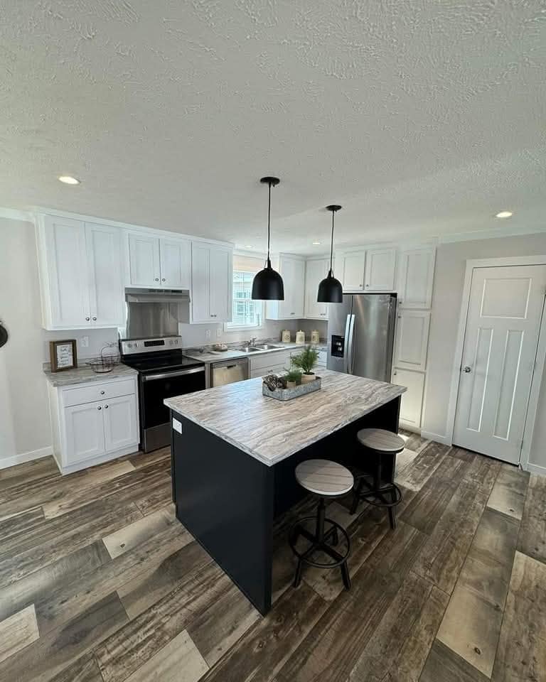 Modern kitchen with wooden floors, a central island with stools, and black pendant lights. White cabinets, stainless steel appliances, bright ambiance.