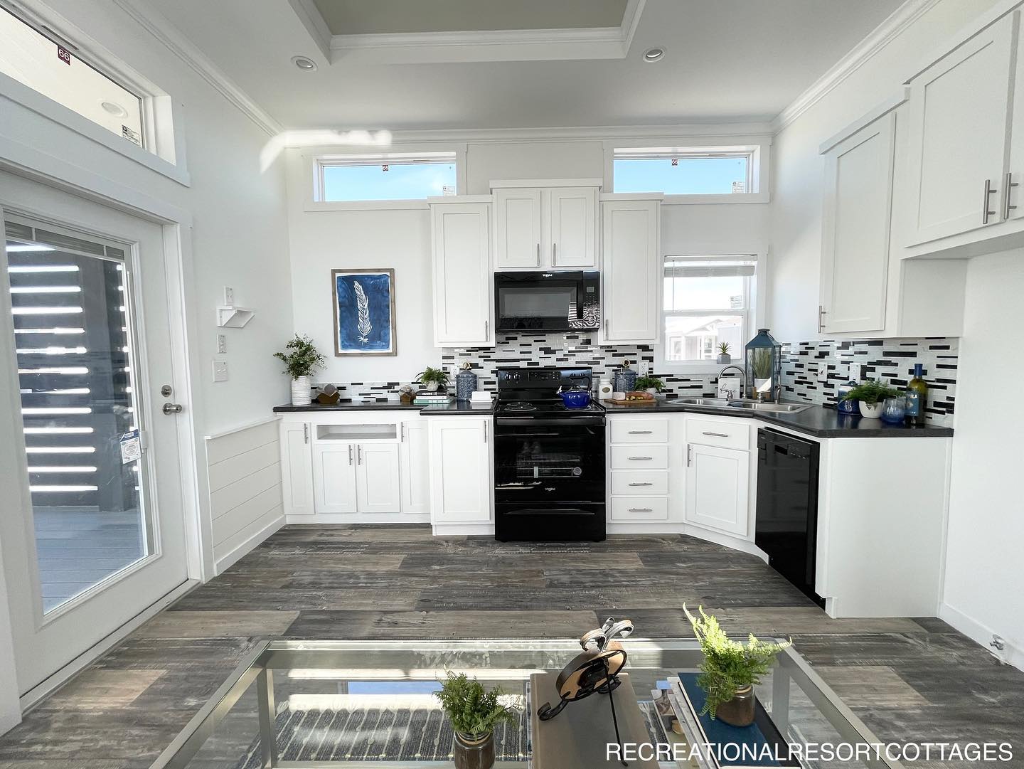 Modern kitchen with white cabinets, black appliances, and a mosaic tile backsplash. Sunlight streams in through high windows, creating a bright, airy feel.