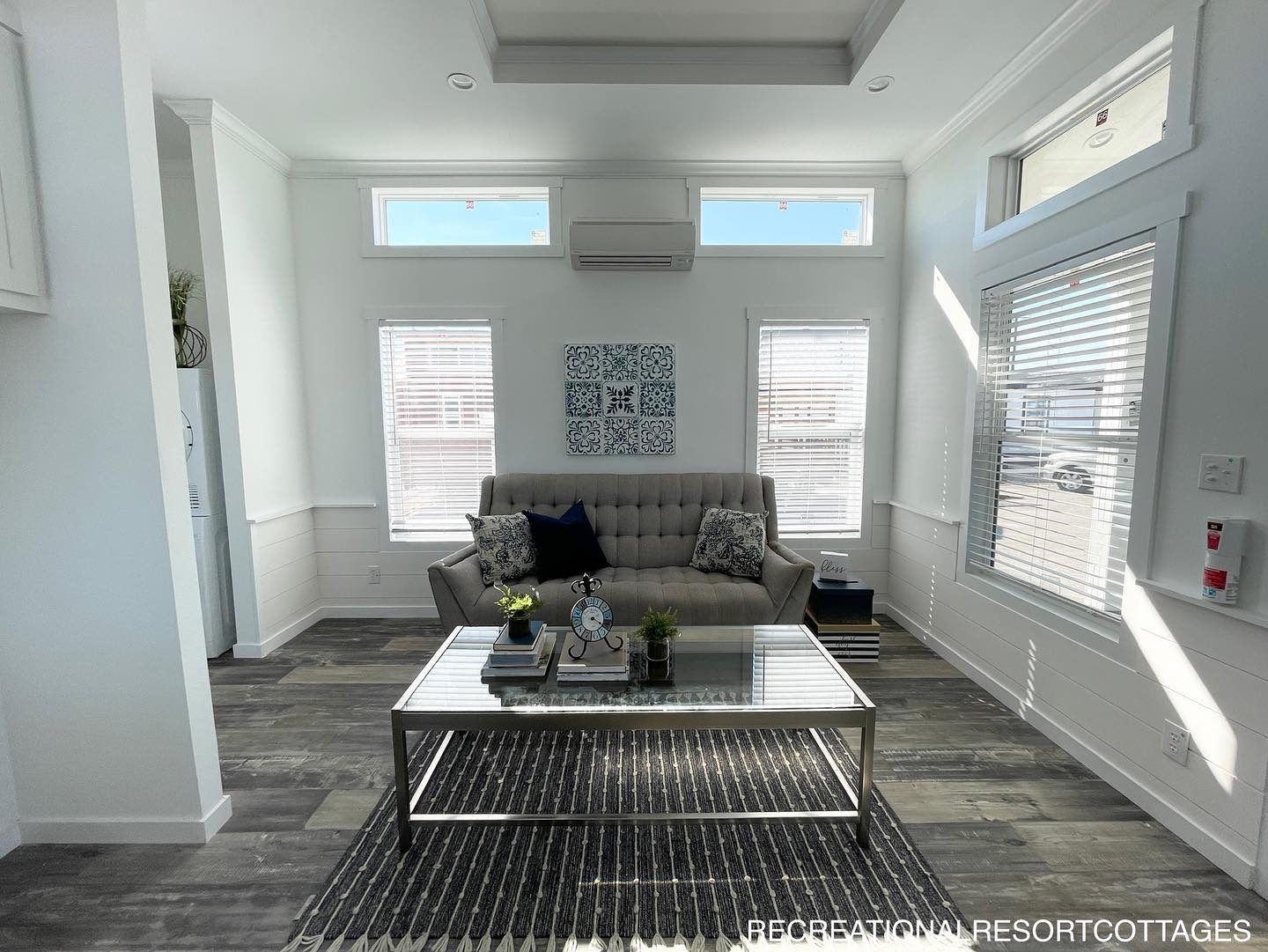 Modern living room with a gray sofa, patterned cushions, and a glass coffee table on a striped rug. Sunlight streams through tall windows, creating a bright, inviting atmosphere.