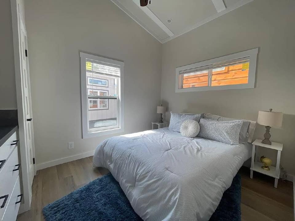 Modern luxury tiny home bedroom featuring a plush white comforter and light blue patterned pillows on a cozy bed, accented by a deep blue shaggy area rug over premium light wood flooring, flanked by white minimalist nightstands with elegant lamps and set against clean grey walls with large bright windows for a sophisticated move-in ready turn-key residential design.