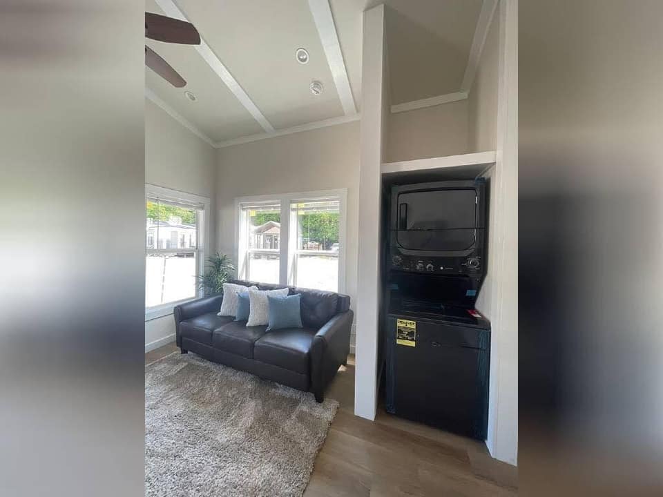 Modern luxury tiny home interior featuring a compact living room with a dark leather sofa, a plush grey area rug, and a built-in laundry nook with a space-saving stacked washer and dryer, showcased under a white vaulted ceiling with recessed lighting for a sophisticated move-in ready turn-key residential design.
