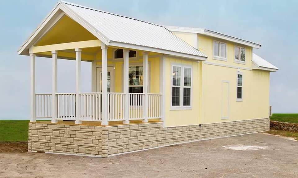 A small, yellow modular house with white trim and a metal roof. It features a cozy front porch with railings and stone foundation, set against a grassy field.