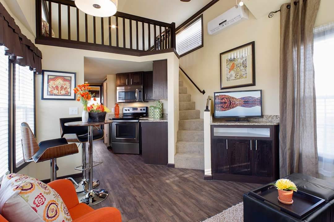 Modern living space with an open loft and staircase. Features dark wood flooring, compact kitchen, vibrant orange chair, and decorative art. Cozy and stylish.