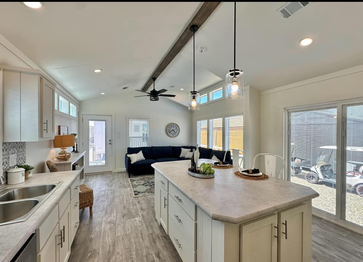 Bright, modern kitchen with white cabinets, island, and pendant lighting. Living area has a navy sofa, large windows, and a wall clock, creating an airy and inviting atmosphere.