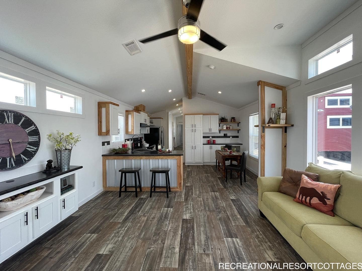 Spacious open-plan living area with modern kitchen, wooden floors, ceiling fan, and large windows. Cozy seating and decorative clock enhance warmth.