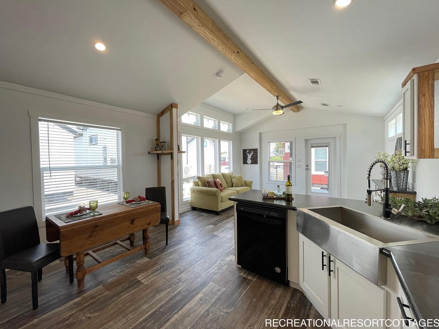 Bright open-plan living space with wooden floors, a kitchen featuring a large sink, and dining area with a rustic table. Cozy sofa under tall windows. Airy and inviting.