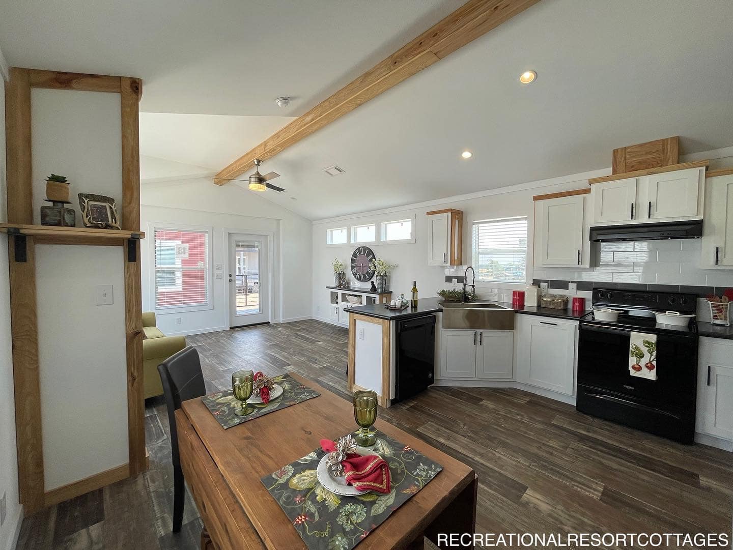 Spacious modern kitchen with white cabinets, black appliances, wooden beams, and a small dining table set for two. Bright, with large windows.
