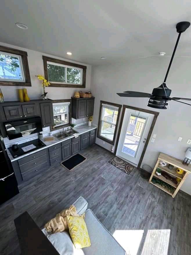 Modern kitchen with gray cabinets, white countertops, and a gray floor. Large windows let in natural light. Cozy seating area and decorative plants.